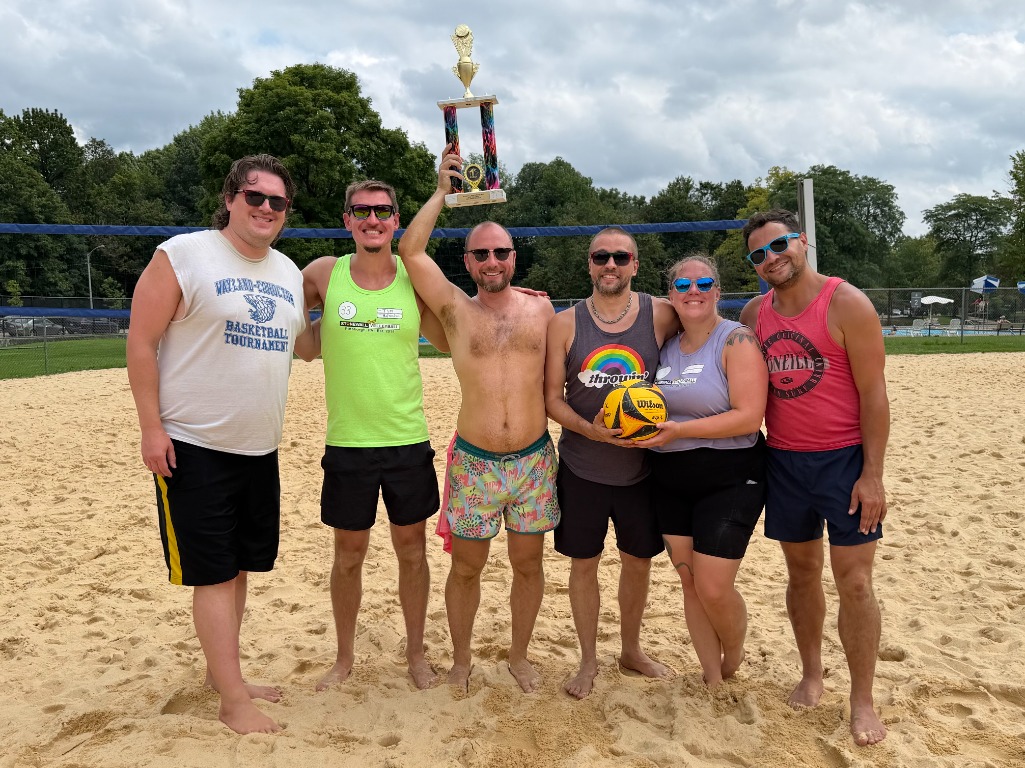 The 2026 Stonewall Volleyball Pittsburgh board together on the sand courts, holding a trophy and a volleyball